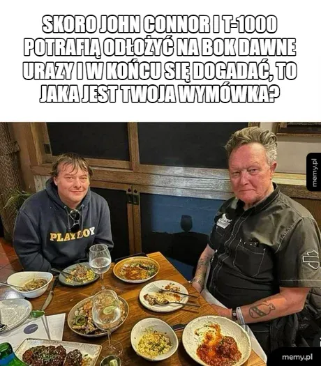 Dwaj mężczyźni siedzą przy stole z jedzeniem, uśmiechając się do kamery.